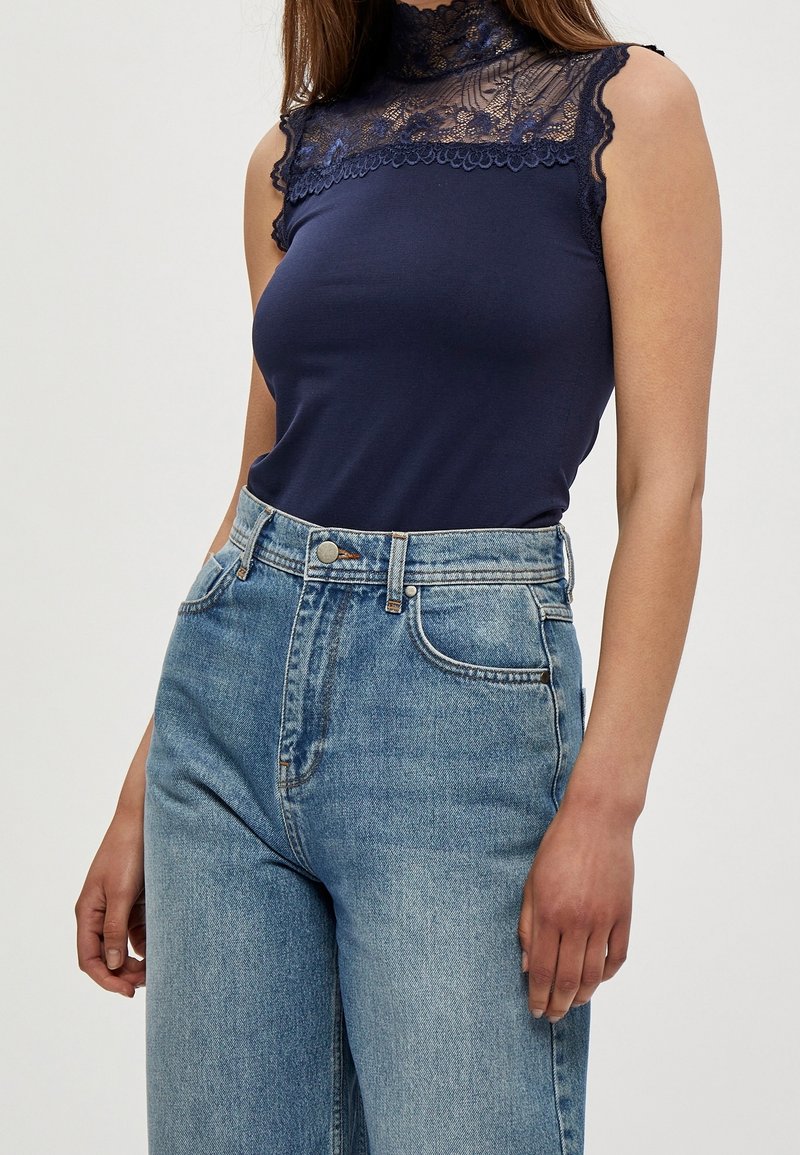 Haut sans manches bleu marine avec un col en dentelle haut, associé à un jean taille haute bleu clair avec une fermeture à boutons et à fermeture éclair classique.