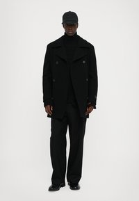 Svart ull peacoat med dubbelknäppt fram, svart ribbad polotröja, svarta byxor och keps. Slät textur och skräddarsydd passform.