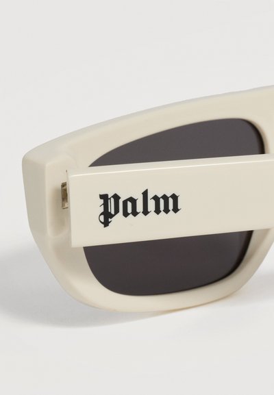 Gros plan sur la branche beige de lunettes de soleil avec le texte noir de style gothique "palm" et une lentille teintée foncée en arrière-plan.
