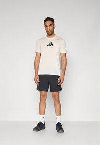 Conjunto desportivo masculino com camisa de manga curta bege clara com logótipo preto da adidas, combinada com calções escuros e sapatilhas de corrida pretas.