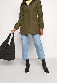 Olijfgroen parka-jack met een capuchon, voorzakken, gecombineerd met lichtblauwe cropped jeans en zwarte enkellaarsjes. Zwarte handtas in de hand.