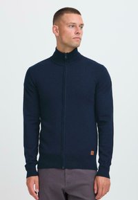 Muškarac kratke kose nosi tamnoplavu duksericu s punim zipom i sive hlače, gleda udesno na jednostavnoj svijetloj pozadini.