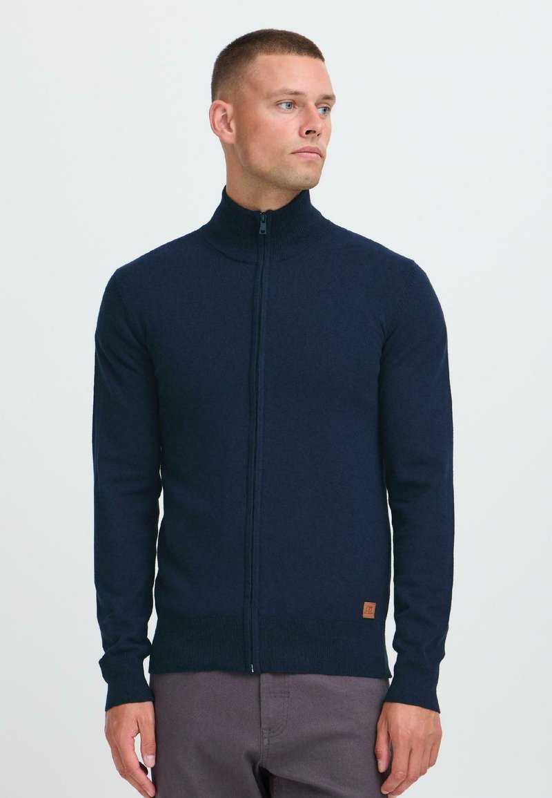 Hombre con cabello corto vestido con un suéter azul marino de cremallera completa y pantalones grises, mirando hacia la derecha contra un fondo claro y sencillo.