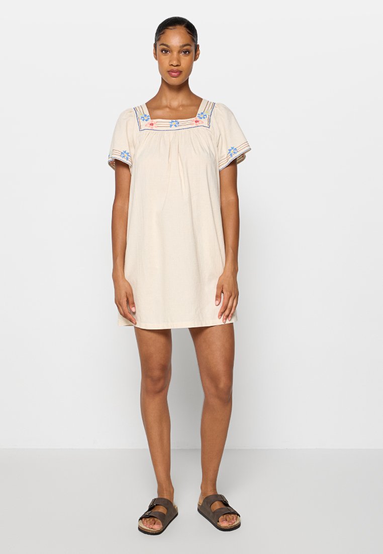 Vrouw staat in een crème-kleurige jurk met korte mouwen, versierd met bloemenborduursel bij de halslijn en mouwen, gecombineerd met donkerbruine sandalen.