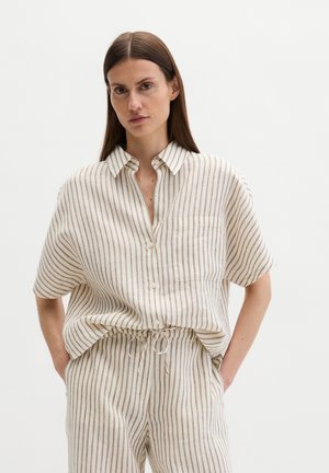 Frau mit langen braunen Haaren, die ein beiges und braunes kurzärmliges Hemd mit vertikalen Streifen sowie passende Hose mit Kordelzug trägt, Hände in den Taschen.