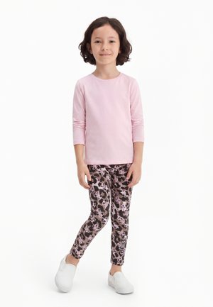 Fată tânără în picioare, purtând un tricou roz deschis cu mâneci lungi, colanți cu modele și logo-uri, și pantofi slip-on albi.