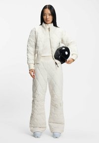 Pikowana biała kurtka puchowa z wysokim kołnierzem, dobrana do szerokich spodni, oba z geometrycznym wzorem i zamkami błyskawicznymi. W zestawie czarny kask.