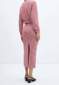 Robe midi à motifs rouges et roses avec manches longues, fente au dos et silhouette ajustée, confectionnée en tissu texturé. Sandales noires à talons carrés visibles.