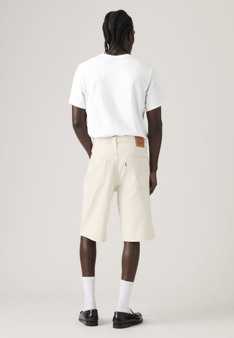 Homme aux cheveux tressés de dos, portant un T-shirt blanc, un short beige jusqu'aux genoux, des chaussettes blanches et des chaussures noires, sur un fond uni.