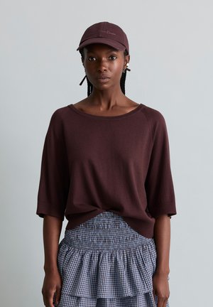 Vrouw die een bordeauxrode top met halve mouwen en pet draagt, gecombineerd met een blauwe en grijze gelaagde geruite rok, staand tegen een effen achtergrond.