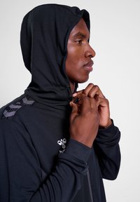 Zwarte hoodie van een gestructureerde stof, slim fit ontwerp en verstelbare koordjes. Beschikt over een logo en gemusterde accenten op de schouders.