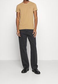 T-shirt beige à manches courtes avec un petit logo sur la poitrine, associé à un pantalon large gris foncé et des chaussures noires.