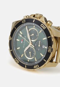 Montre en métal doré avec une lunette noire, un cadran vert et des sous-cadrans chronographes. Présente des marqueurs dorés et une fenêtre de date à 4 heures.