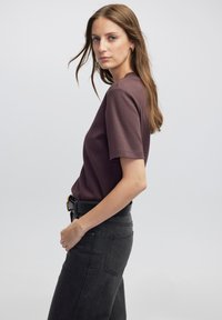Camisa marrón de manga corta combinada con jeans negros de cintura alta. El look incluye un cinturón negro con una hebilla dorada, destacando un estilo casual.