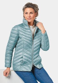 Giacca puffer azzurro chiaro con cuciture a chevron, colletto alto e chiusura con zip. Tessuto morbido e tasche laterali. Indossata sopra una maglietta bianca.