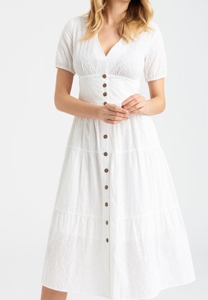 Mujer vistiendo un vestido midi blanco con bordado, de mangas cortas y botones marrones en la parte delantera, de pie con las manos suavemente entrelazadas.