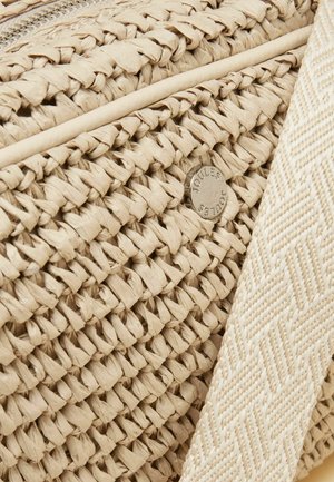 Detalje af beigefarvet flettet stråtaske med en tekstureret stofrem og en lille rund metal-logo-mærkat ætset med "Joules."