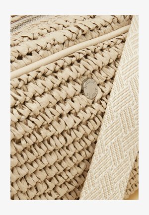 Detalje af beigefarvet flettet stråtaske med en tekstureret stofrem og en lille rund metal-logo-mærkat ætset med "Joules."