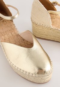 Guldig metallic kilklack med en texturerad yta, öppen tå och övre del i flätat material. Har en sula av juterrep och justerbar rem.