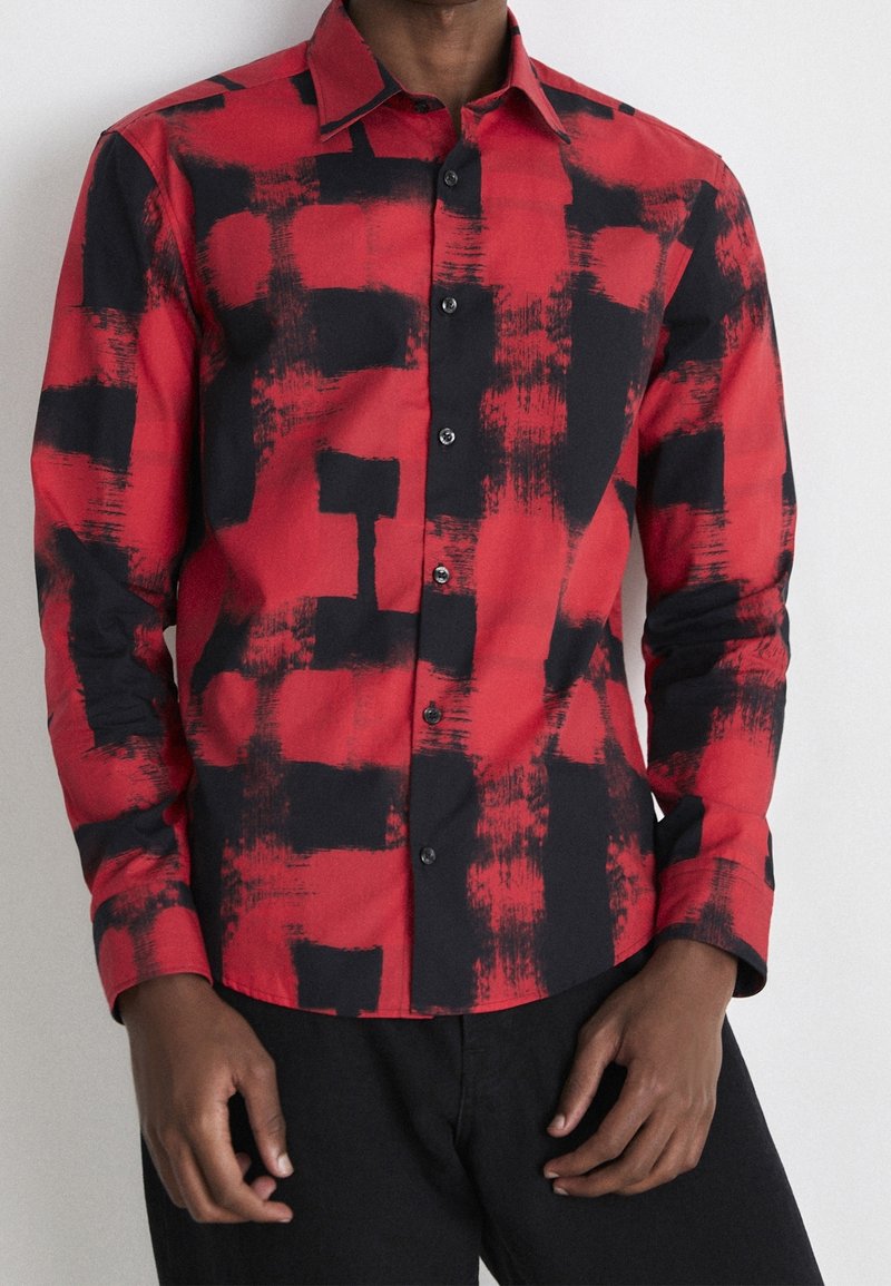 Homme portant une chemise rouge à boutons avec des motifs abstraits audacieux en coups de pinceau noirs, associée à un pantalon noir, debout devant un fond uni.