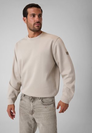 Mand iført en beige langærmet sweatshirt og lysegrå jeans, står op ad en ensartet lysegrå baggrund og ser mod venstre.