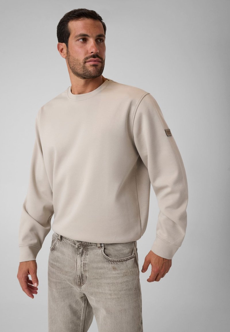 Homme portant un sweat-shirt beige à manches longues et un jean gris clair, debout devant un fond uni gris clair, regardant vers sa gauche.