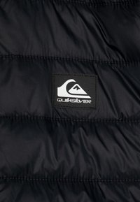 Svart puffjacka med quiltad textur. Har en rektangulär logotypmärke med vit text och logotypdesign.