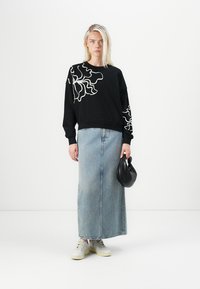 Felpa nera con design ondulato bianco, abbinata a una gonna maxi di jeans azzurri. Il modello tiene una borsa nera indossando sneakers bianche.