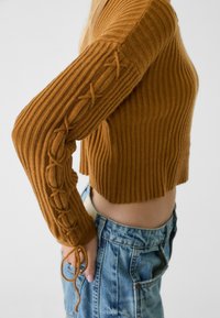 Brązowy sweter z prążkowanej dzianiny z długimi rękawami, z ozdobnym sznurowaniem wzdłuż lewego ramienia i skróconym dołem. Noszony z spodniami z wysokim stanem.