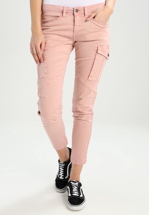 Osoba nosząca różowe skrócone spodnie bojówki z bocznymi kieszeniami oraz czarne sznurowane trampki na jasnym tle.