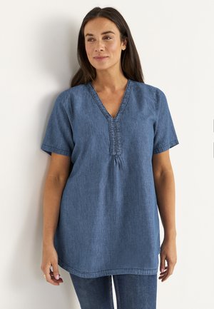 Frau, die eine blaue kurzärmlige Denim-Tunika mit V-Ausschnitt und Rüschensaum trägt, kombiniert mit dunkelblauen Jeans, die vor einer weißen Wand steht.