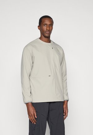 Veste essentielle gris clair, texture douce, encolure ronde, manches longues, avec une petite poche avant et un design minimaliste.