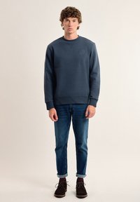 Sudadera azul oscuro con cuello redondo y logo bordado en el pecho, combinada con jeans azul claro y zapatos marrones.
