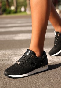 Svarta stickade sneakers med rund tå, textilstruktur, metalliska öljetter och en vit sula med guldaccent på hälen.