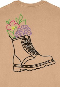 Chemise beige ornée d'une botte noire brodée avec des détails en dentelle, surmontée de fleurs colorées en rose, violet et vert.