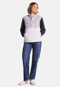 Fleece pullover in light gray and navy with a high collar, snap buttons, and front pockets, paired with blue jeans and sneakers.