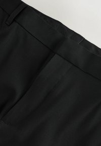 Melnas pielāgotas bikses ar gludu tekstūru, plakanu priekšpusi un slīpām kabatām. Jostasvieta ir eleganta ar smalkiem šuvju elementiem.