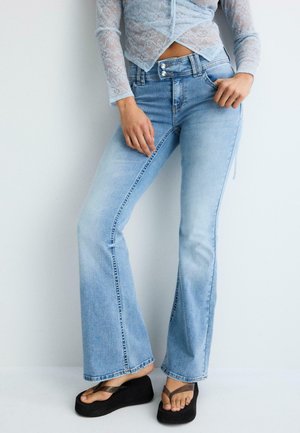 Person iført lyseblå vide jeans, gennemsigtig hvid blondebluse med lange ærmer og sorte plateau-sandaler mod en ensfarvet baggrund.