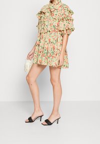 Frau, die ein kurzes, blütenmusteredes Kleid mit Rüschensäumen trägt, eine kleine Perlenhandtasche hält und schwarze, hochhackige Sandalen trägt.