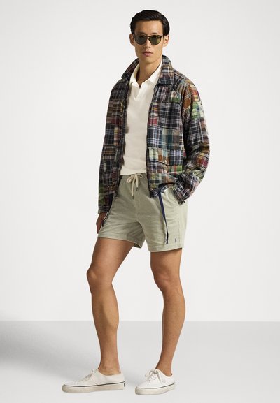 Men's plaid jacket in multicolor fabric, cream polo shirt, light green corduroy shorts, and white sneakers with laces. Round sunglasses.
