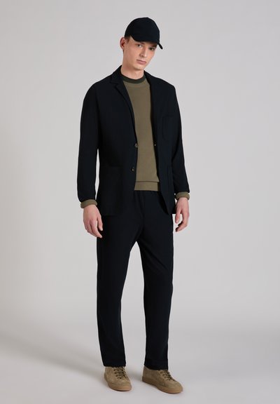 Black tailored blazer over a green sweater, paired with dark trousers and tan sneakers. Features include a chest pocket and button details.