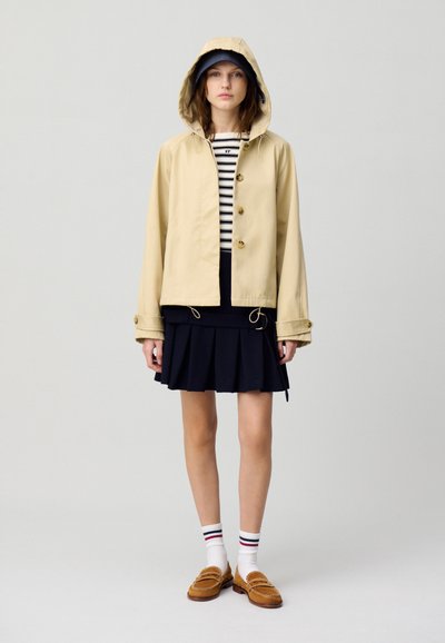 Veste de pluie beige avec capuche, ornée de boutons dorés, portée sur une jupe plissée noire et un t-shirt rayé. Associée à des mocassins marron et des chaussettes blanches.