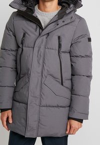 Giacca a vento grigia con zipper frontale, due tasche anteriori e cappuccio rimovibile. Presenta imbottitura cucita e dettagli neri.