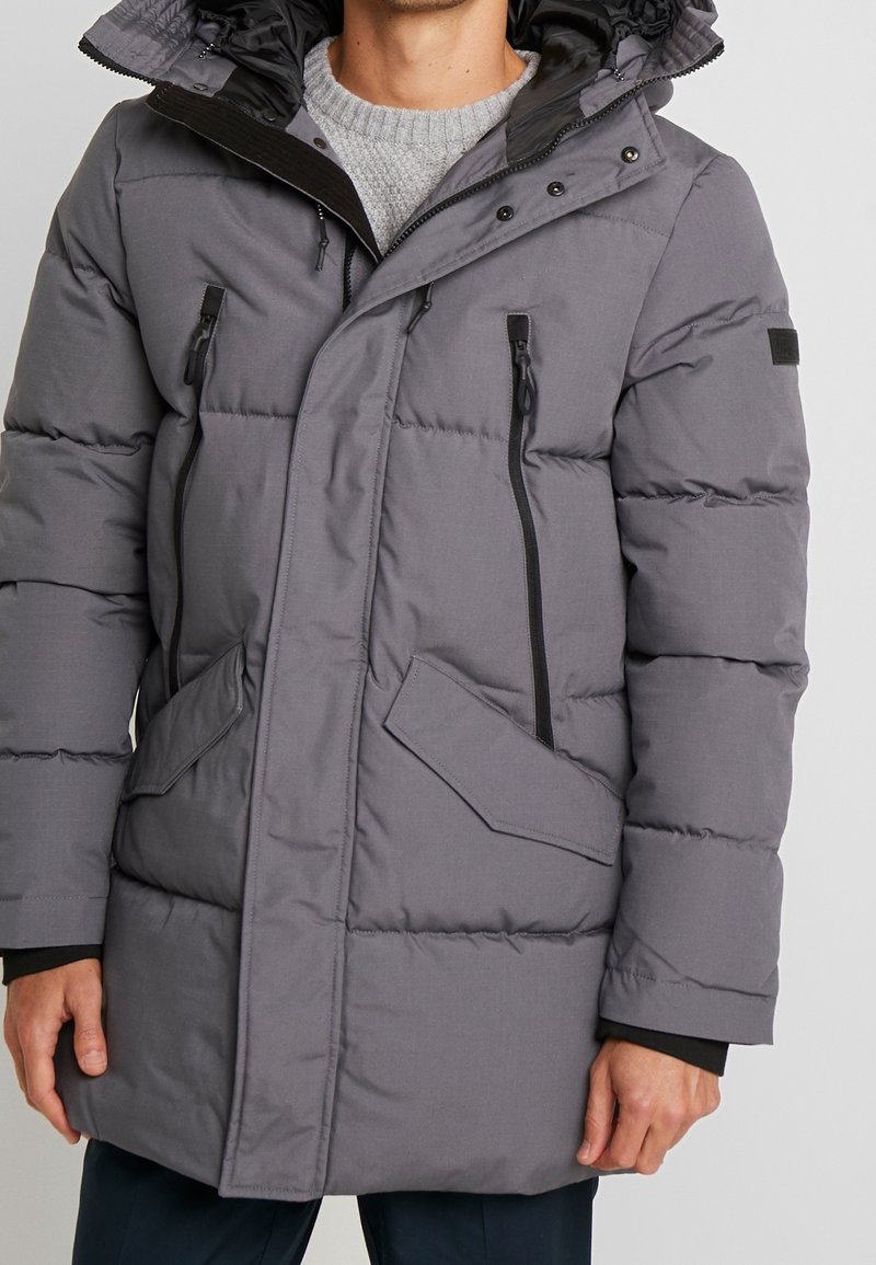 Giacca a vento grigia con zipper frontale, due tasche anteriori e cappuccio rimovibile. Presenta imbottitura cucita e dettagli neri.