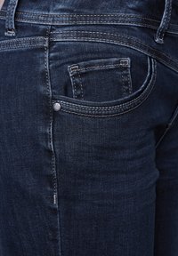Un jean en denim bleu foncé présente une coupe ajustée, des coutures distinctives et une petite poche arrière avec un accent métallique. Surface en tissu texturé.