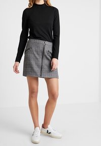 Svart ribbad långärmad topp i kombination med en grå rutig minikjol som har en dragkedja och dekorativ hårdvara, avslutad med vita sneakers.