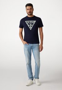 Navy blue t-shirt with a large triangular logo in light colors, paired with light blue jeans and white sneakers. Casual fit and soft texture.