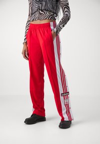 Calças de atletismo vermelhas com riscas laterais brancas, botões de pressão, cós elástico e patch do logótipo da Adidas. Feitas de um material liso e leve.