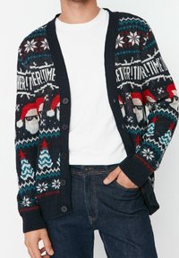 Cardigan blu navy con motivi natalizi, tra cui volti di Babbo Natale, alberi di Natale, fiocchi di neve e la parola "EVERYTIME", indossato sopra una camicia bianca e jeans.