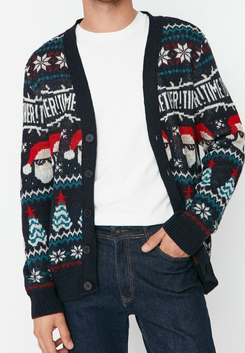 Cardigan blu navy con motivi natalizi, tra cui volti di Babbo Natale, alberi di Natale, fiocchi di neve e la parola "EVERYTIME", indossato sopra una camicia bianca e jeans.